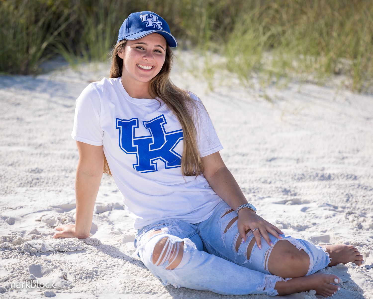 A University of Kentucky fan in a white team shirt and distressed jeans poses on a sandy beach.