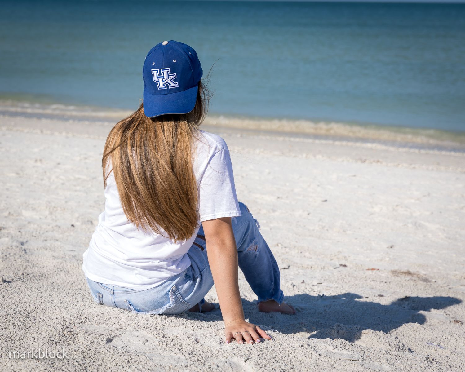 Someone wearing a University of Kentucky baseball cap sits on a sandy beach looking out at blue ocean waters.