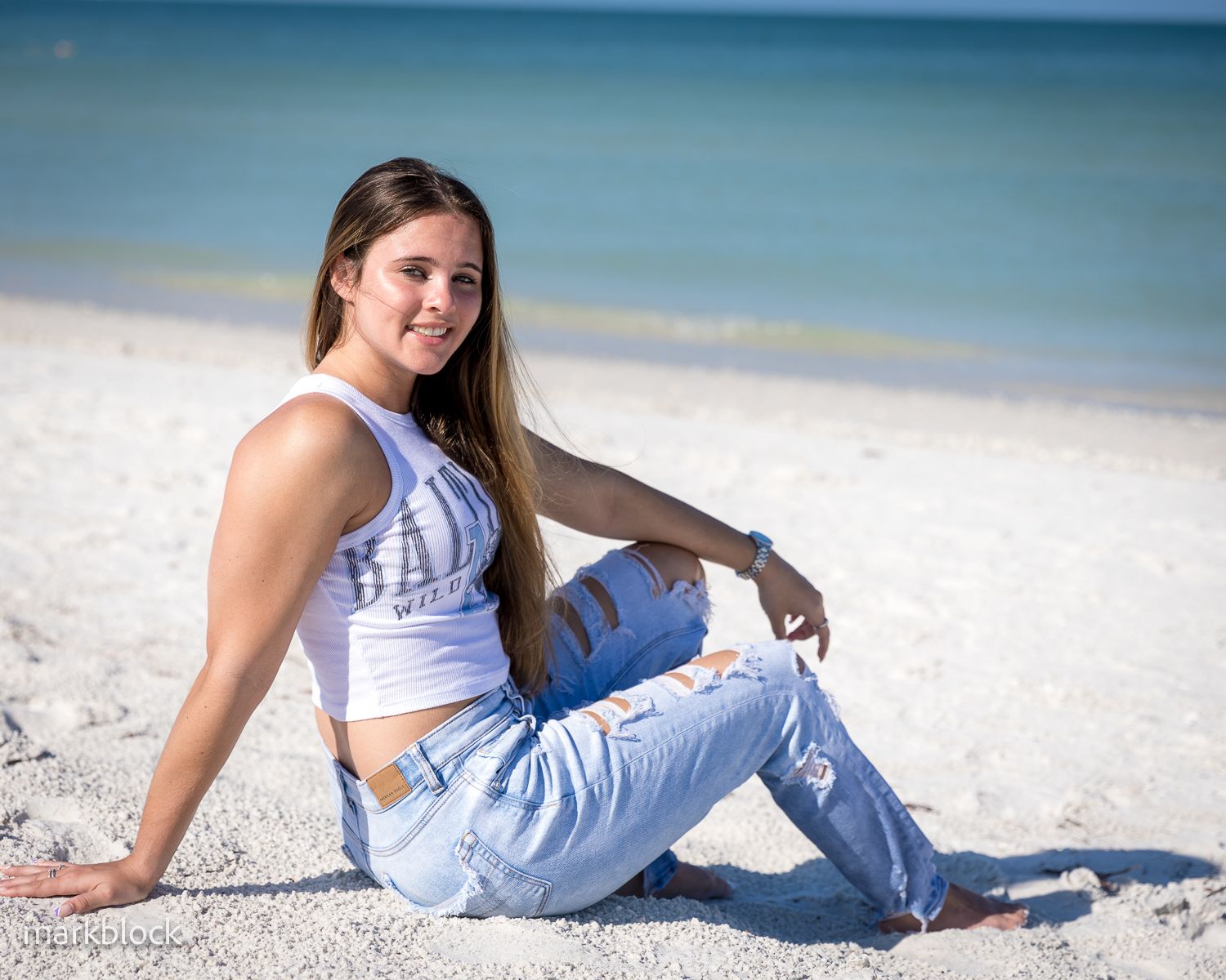 A beachy lifestyle photo of someone in ripped jeans and a white crop top sitting on pristine white sand.