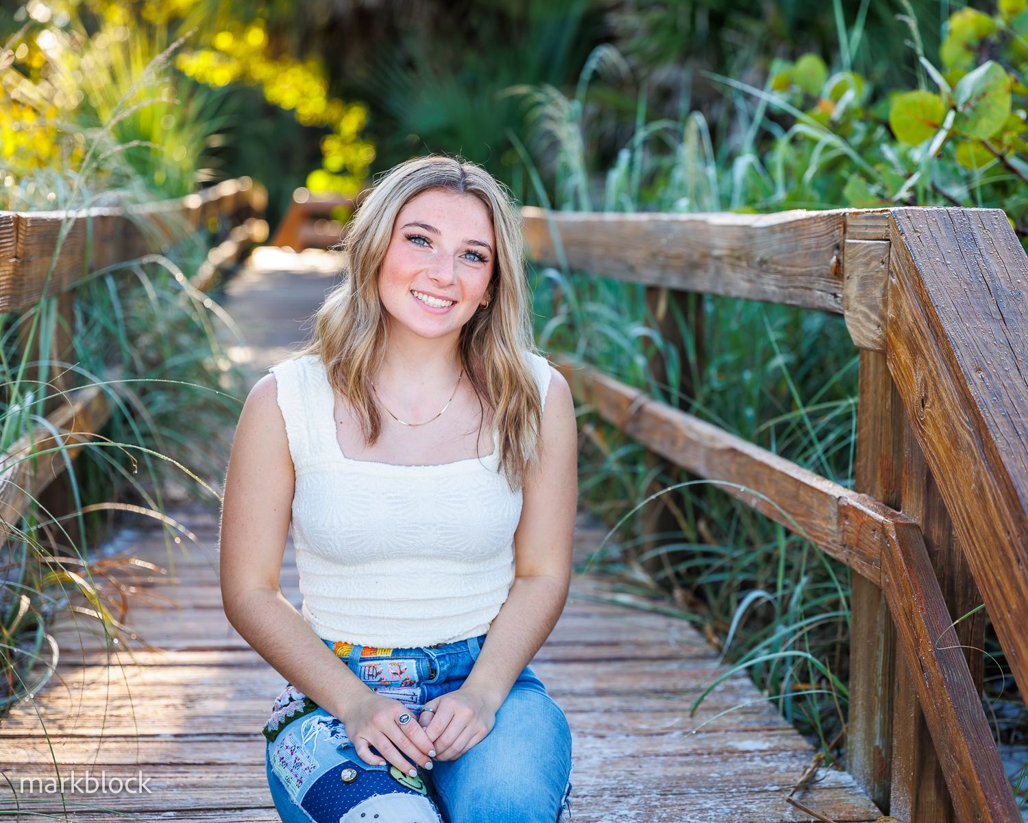 A person in a white top and jeans sits smiling on a wooden boardwalk surrounded by vibrant yellow wildflowers and greenery.
