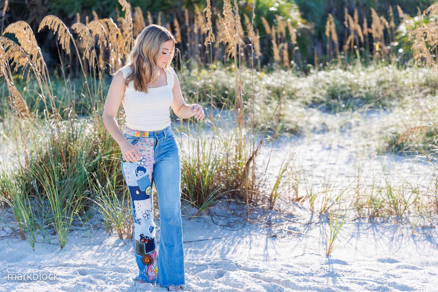 Beachside figure in white top and embellished flare jeans poses among coastal grasses on sandy dunes.