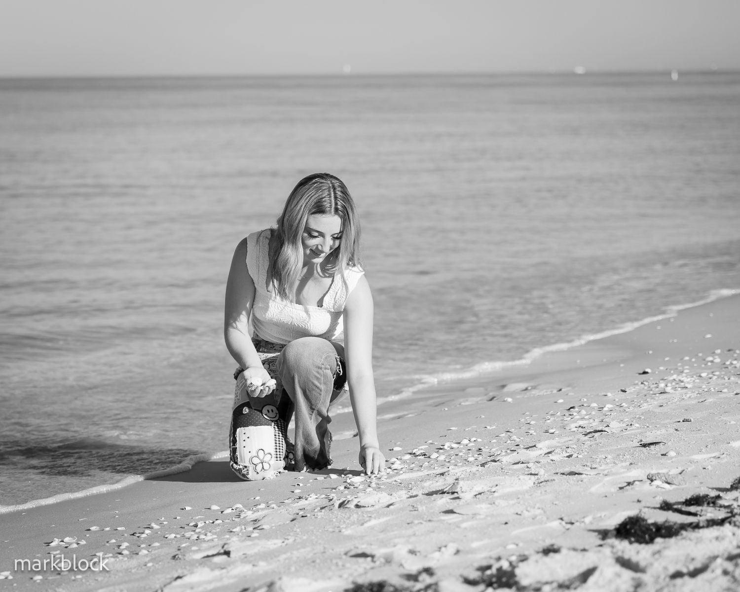 A contemplative figure crouches on a serene black and white beach shoreline, collecting shells by the ocean.