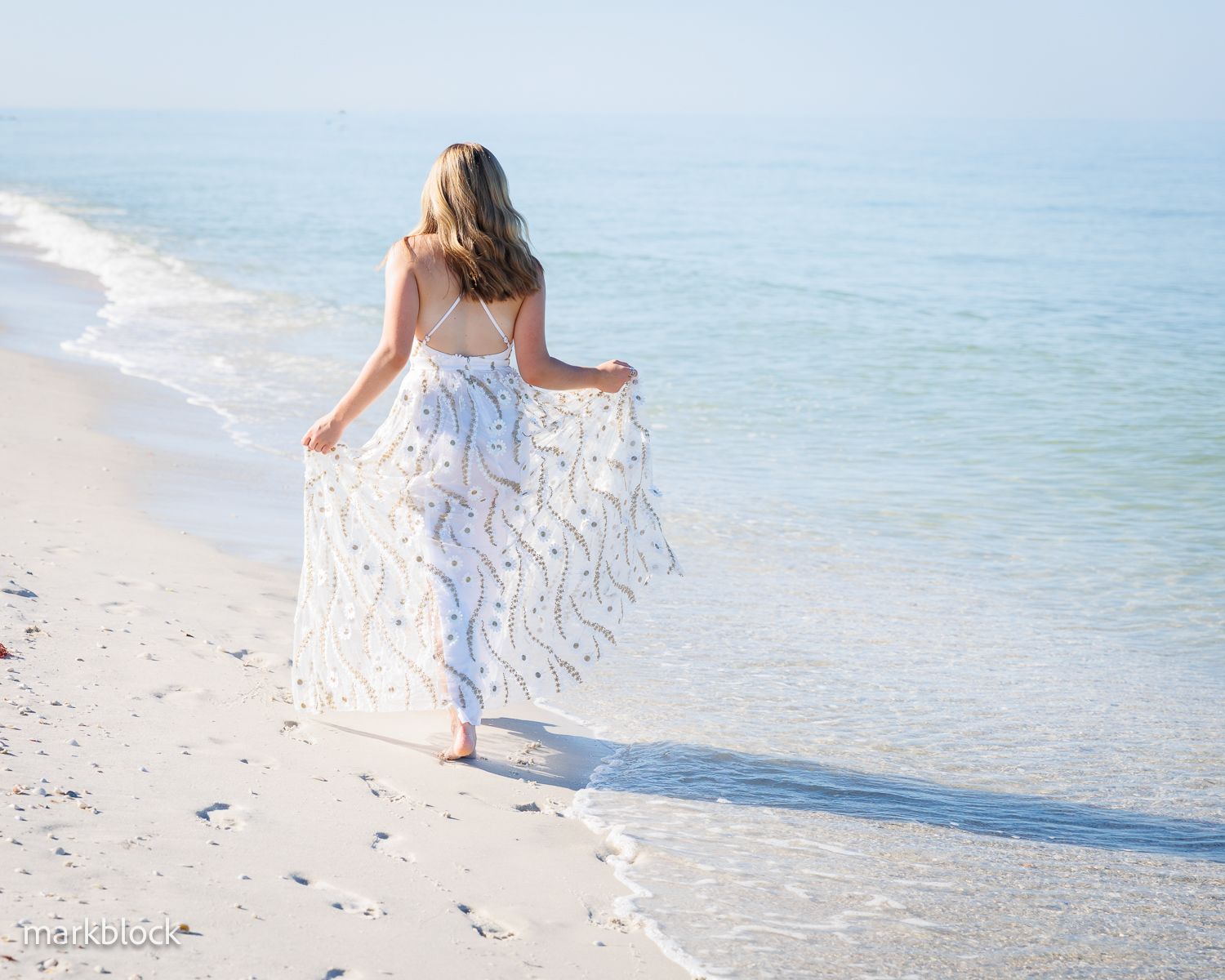 A person in a white flowing dress walks along a pristine sandy beach with turquoise ocean waters in the background.