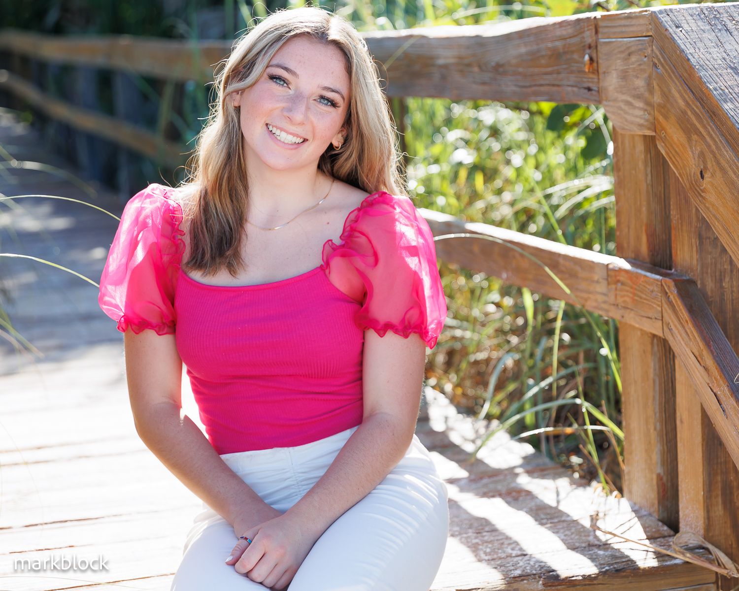Someone in a bright pink top and white pants sits smiling on wooden steps outdoors on a sunny day.