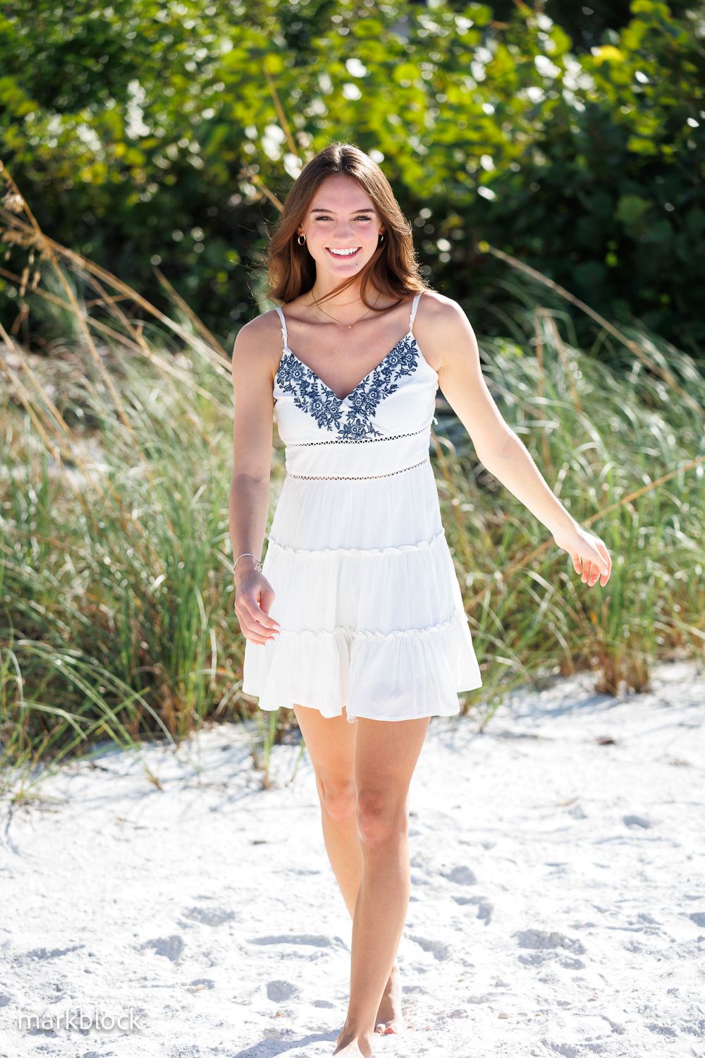 A white summer dress with navy blue embroidery details being modeled on a sandy beach with lush greenery in background.