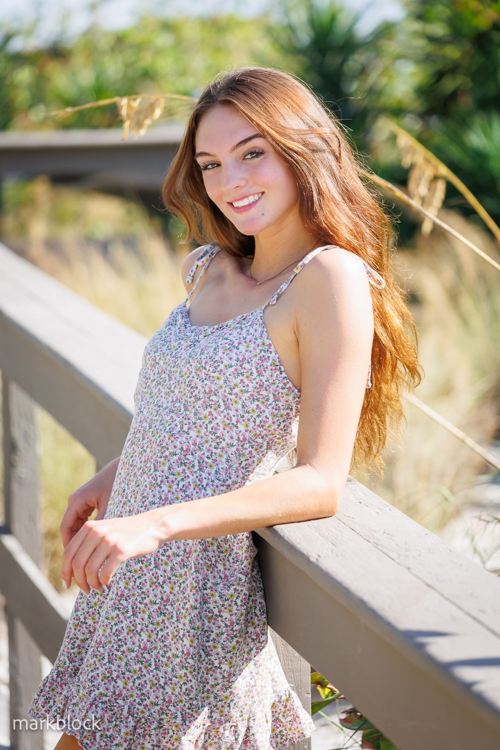 A floral dress portrait on a sunny boardwalk with natural lighting and coastal vegetation in the background.