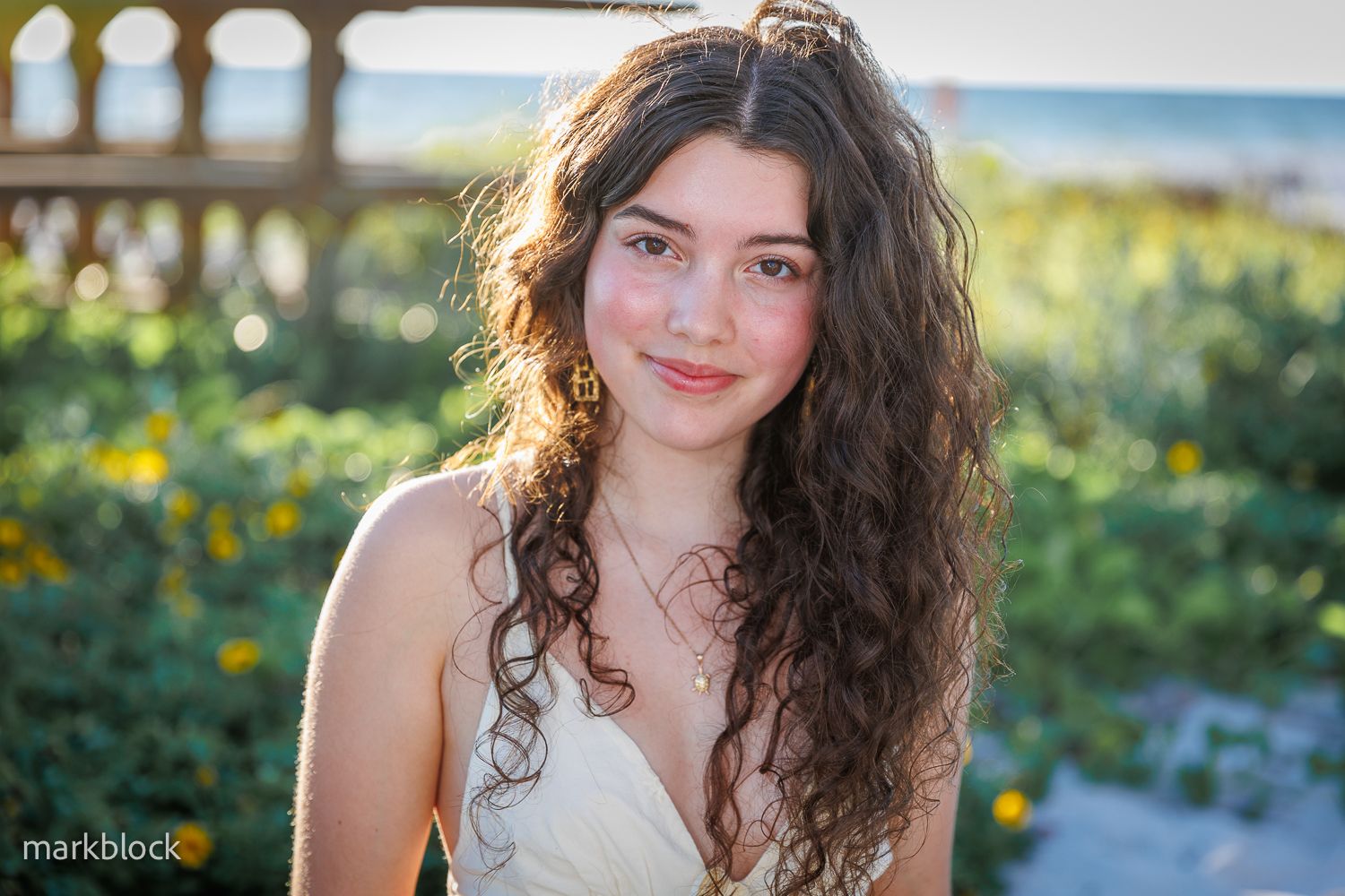 A person with long curly dark hair and a white top poses outdoors in a garden setting during golden hour.