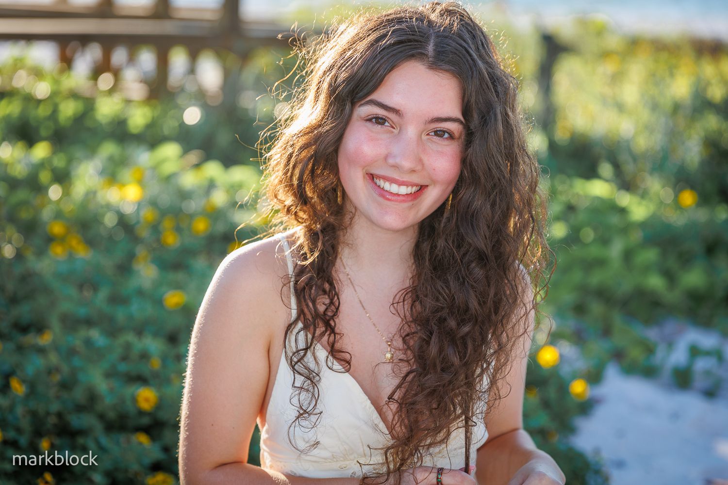 A portrait of someone with long curly brown hair wearing a white top in a sunny garden setting.