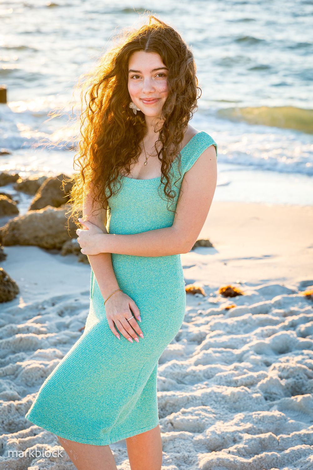 Woman with long curly dark hair wearing a turquoise dress poses on a sunny beach at golden hour.