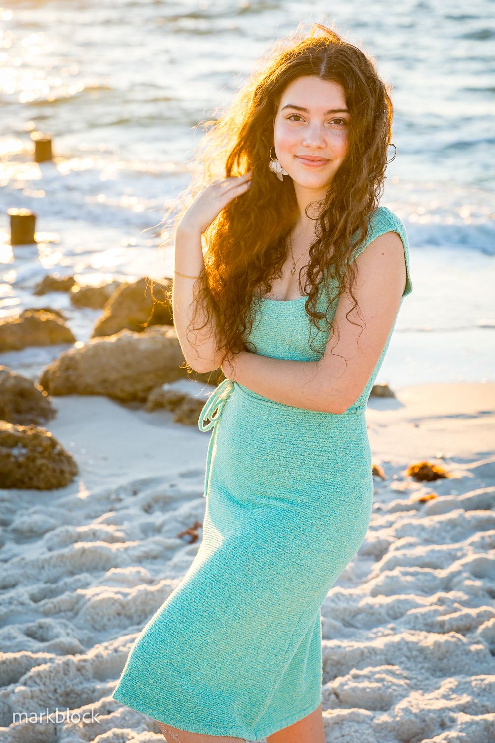 A person in a turquoise summer dress poses on a sandy beach during golden hour with waves and rocks in the background.