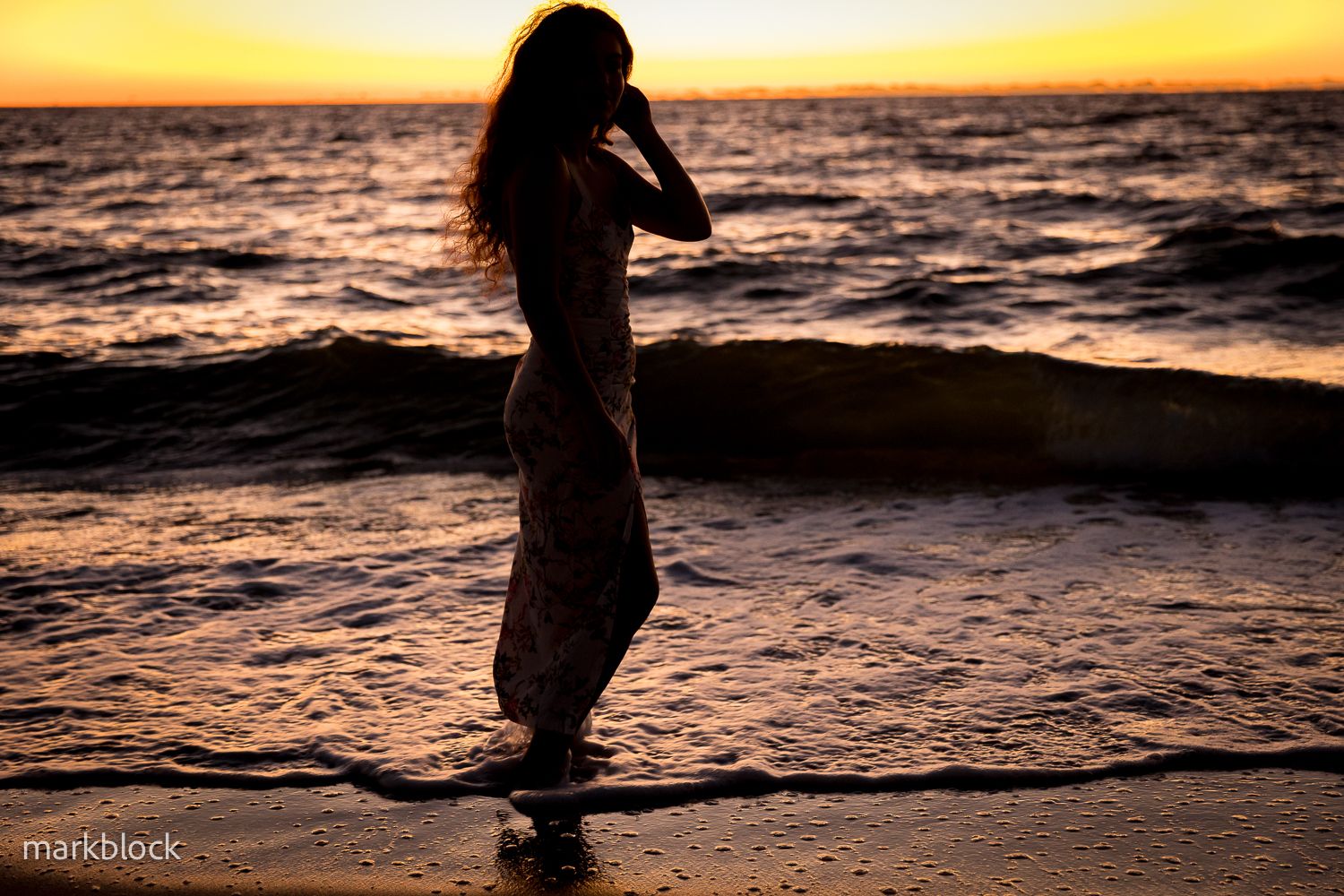 A silhouetted figure stands in shallow ocean waves against a vibrant golden sunset sky.