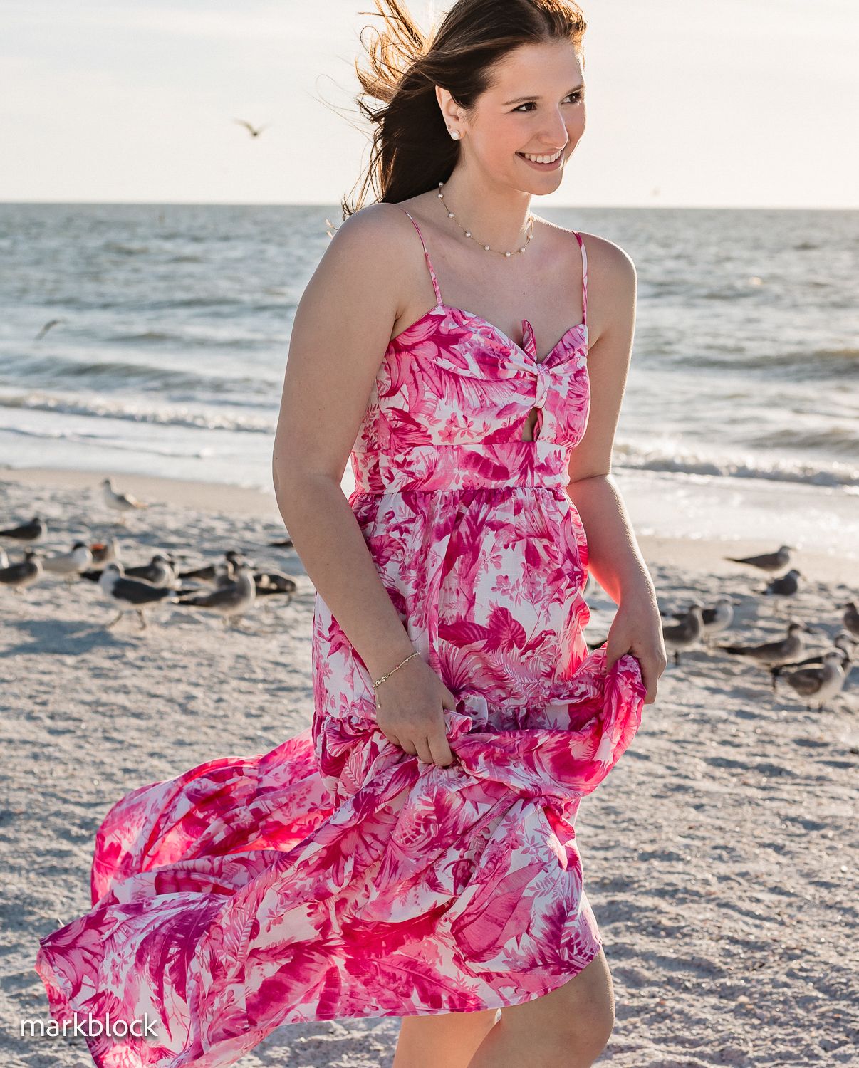 A person in a flowing pink and white floral sundress walks along a beach at sunset with hair blowing in the wind.