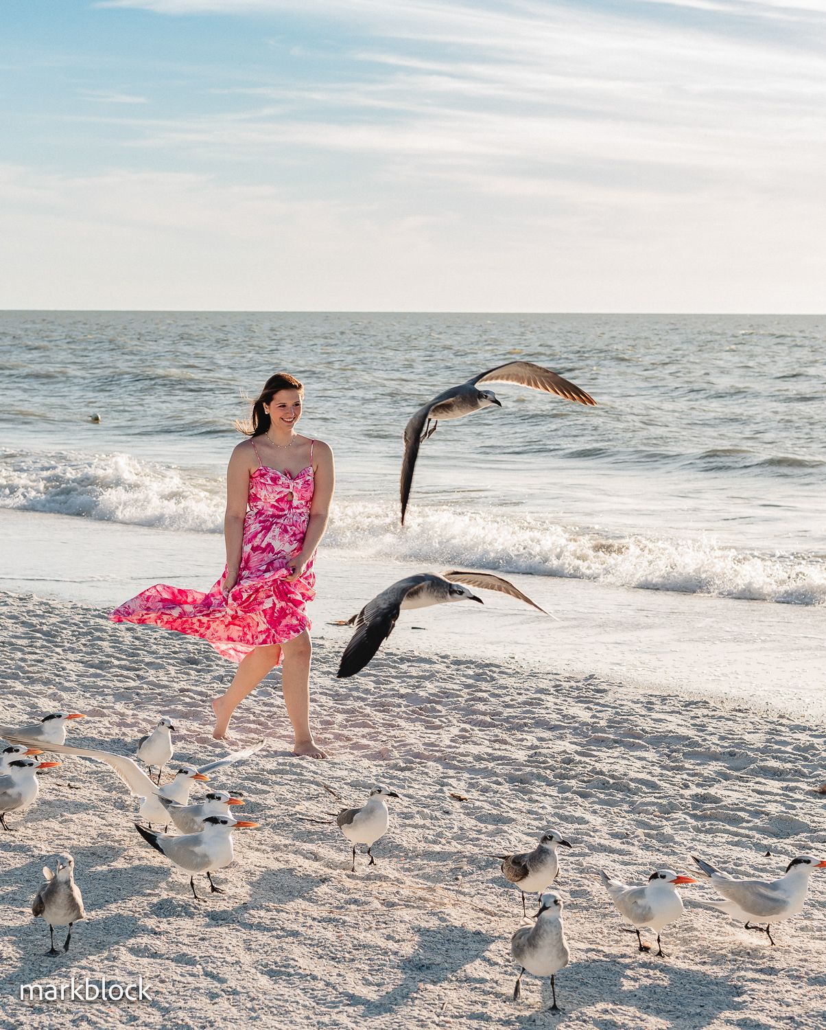 A person in a pink floral dress walks along a sandy beach while seagulls fly and gather around them at sunset.
