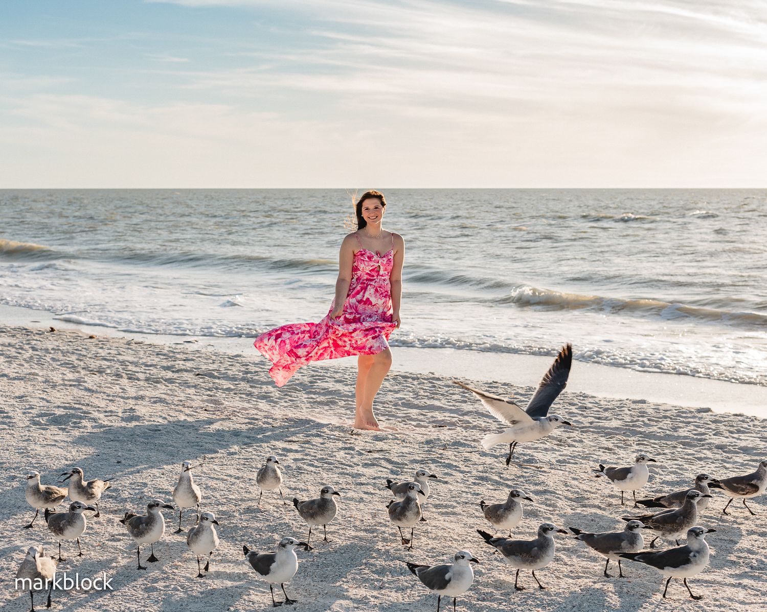 A person in a pink floral dress stands on a beach at sunset surrounded by seabirds on the sand.