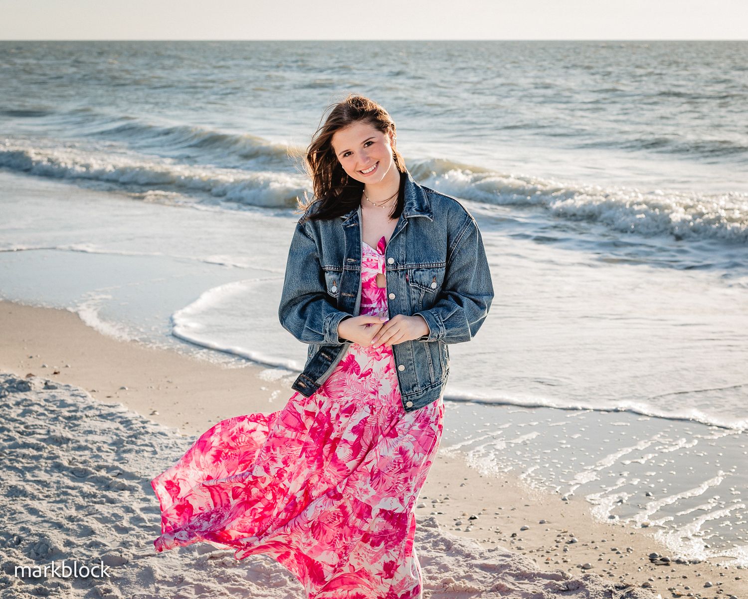 Someone wearing a pink floral dress and denim jacket poses on a beach with waves crashing in the background.