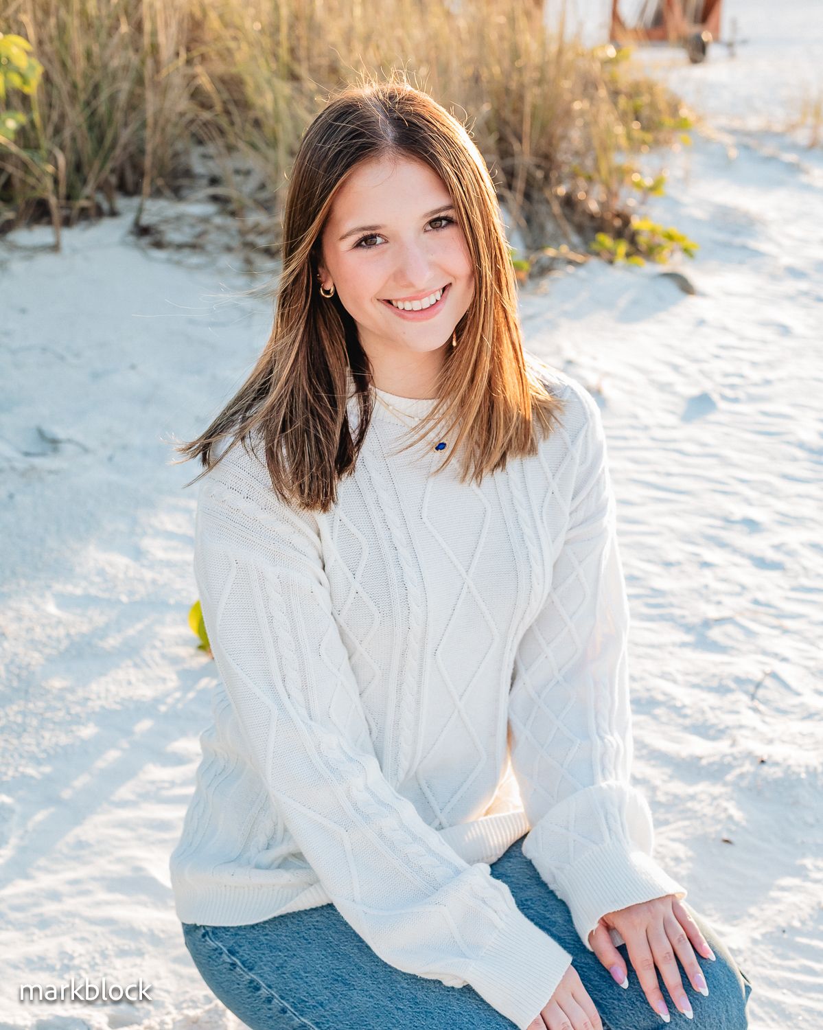 Person in white cable knit sweater and jeans sitting in snowy outdoor setting with natural sunlight.