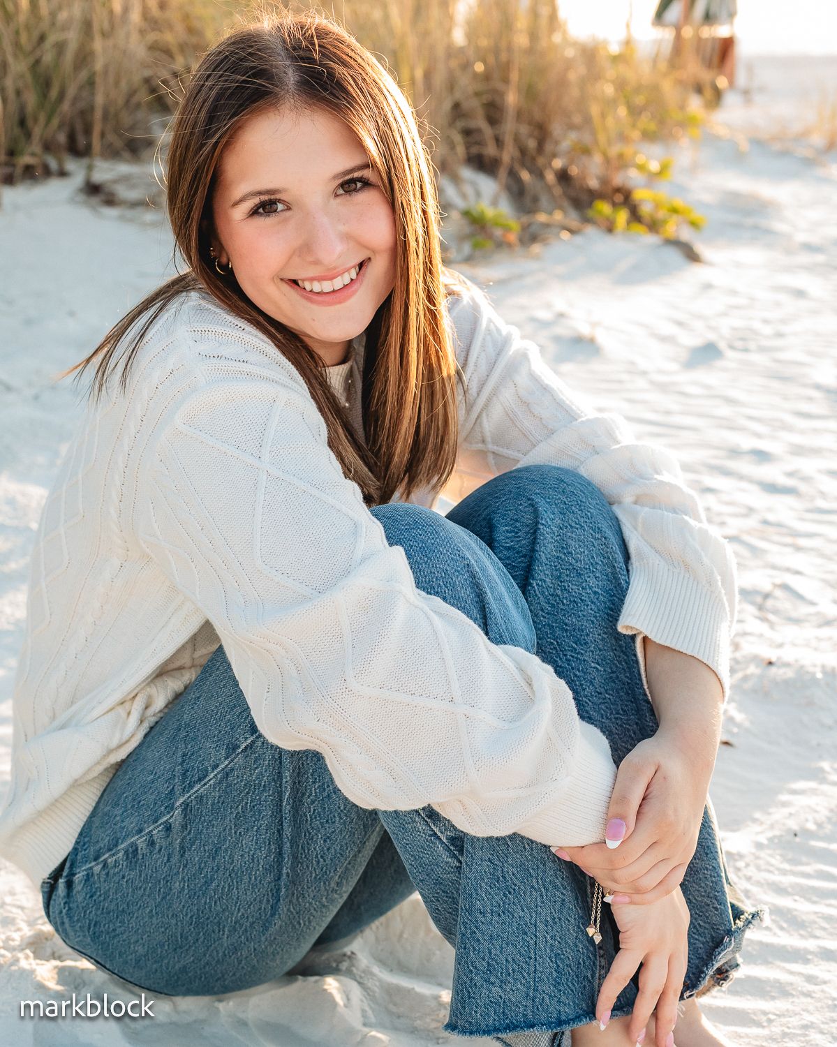 Person in cozy white sweater and blue jeans poses casually on snowy ground with tall grass in background.