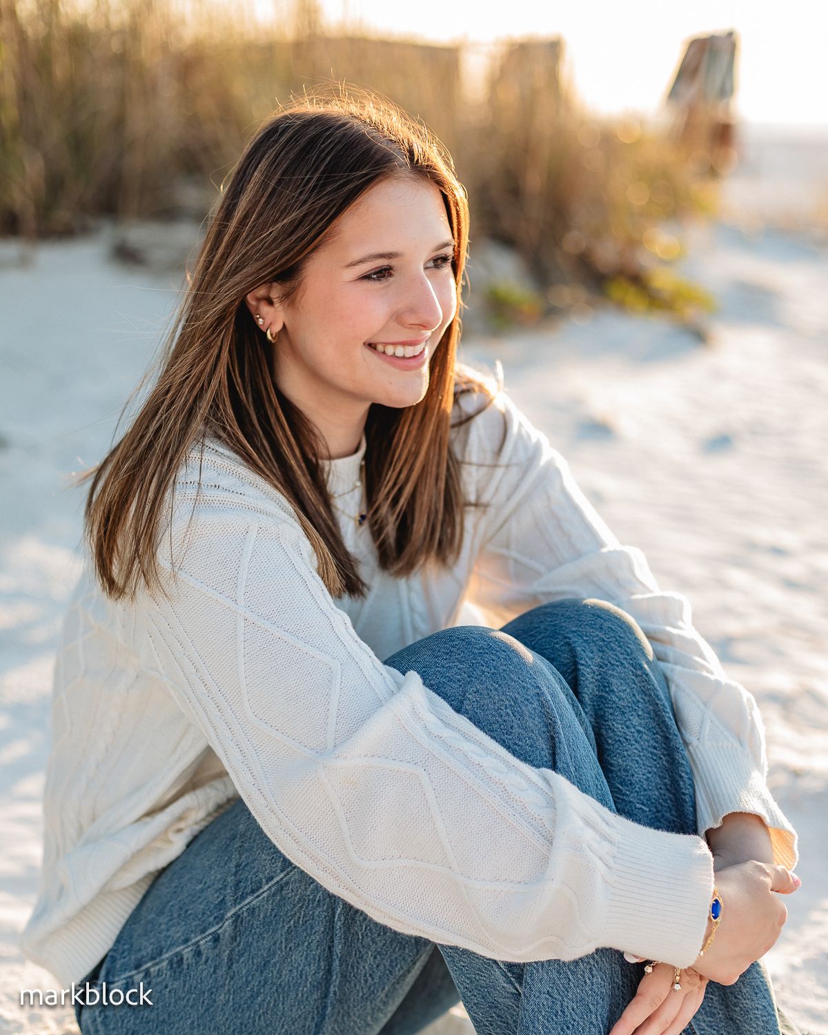 Person in white sweater and jeans sitting on sand at beach during golden hour sunset with natural lighting.
