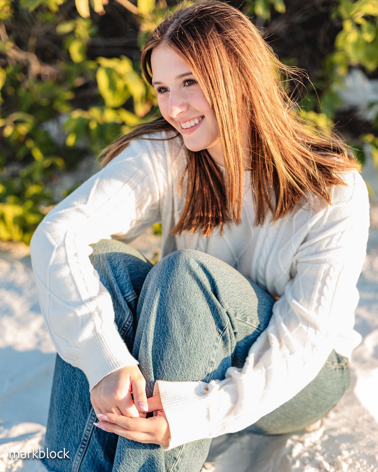 Young person in white sweater and jeans sitting outdoors with golden sunlight and green foliage in background.