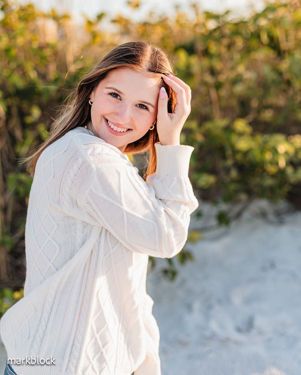 Young person wearing a cozy white cable knit sweater smiles warmly in soft outdoor lighting at sunset.