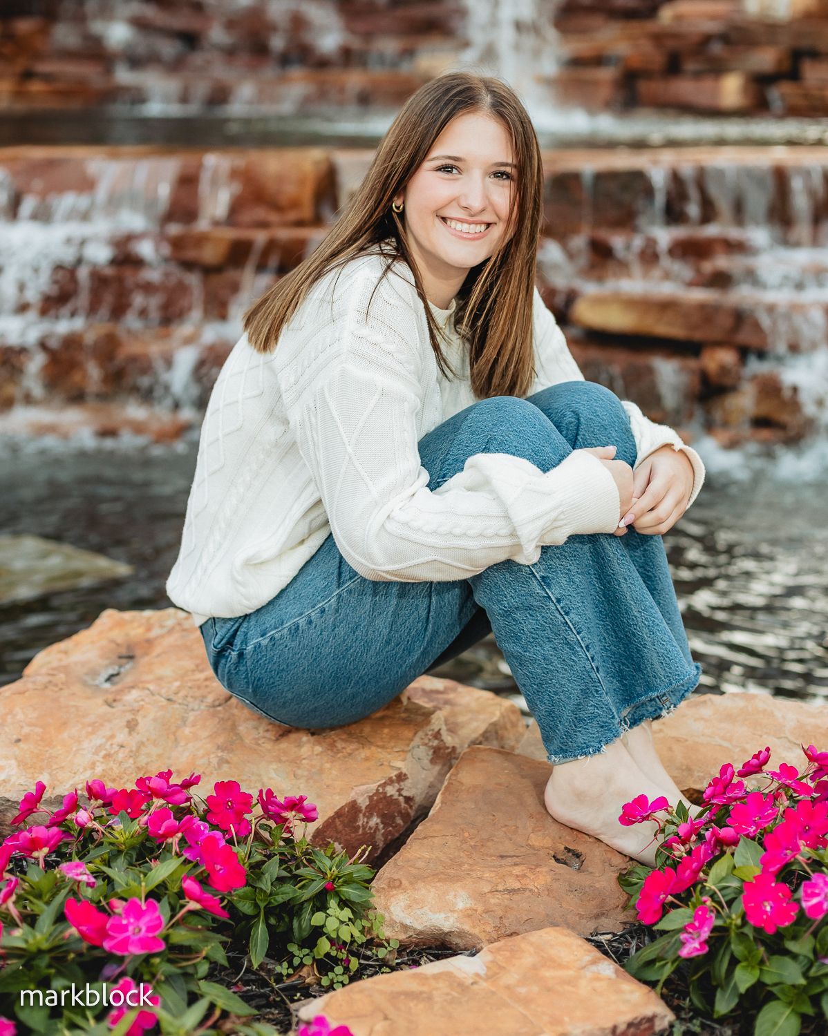Casual portrait of a young person in white sweater and jeans sitting on brick steps with pink flowers beside them.