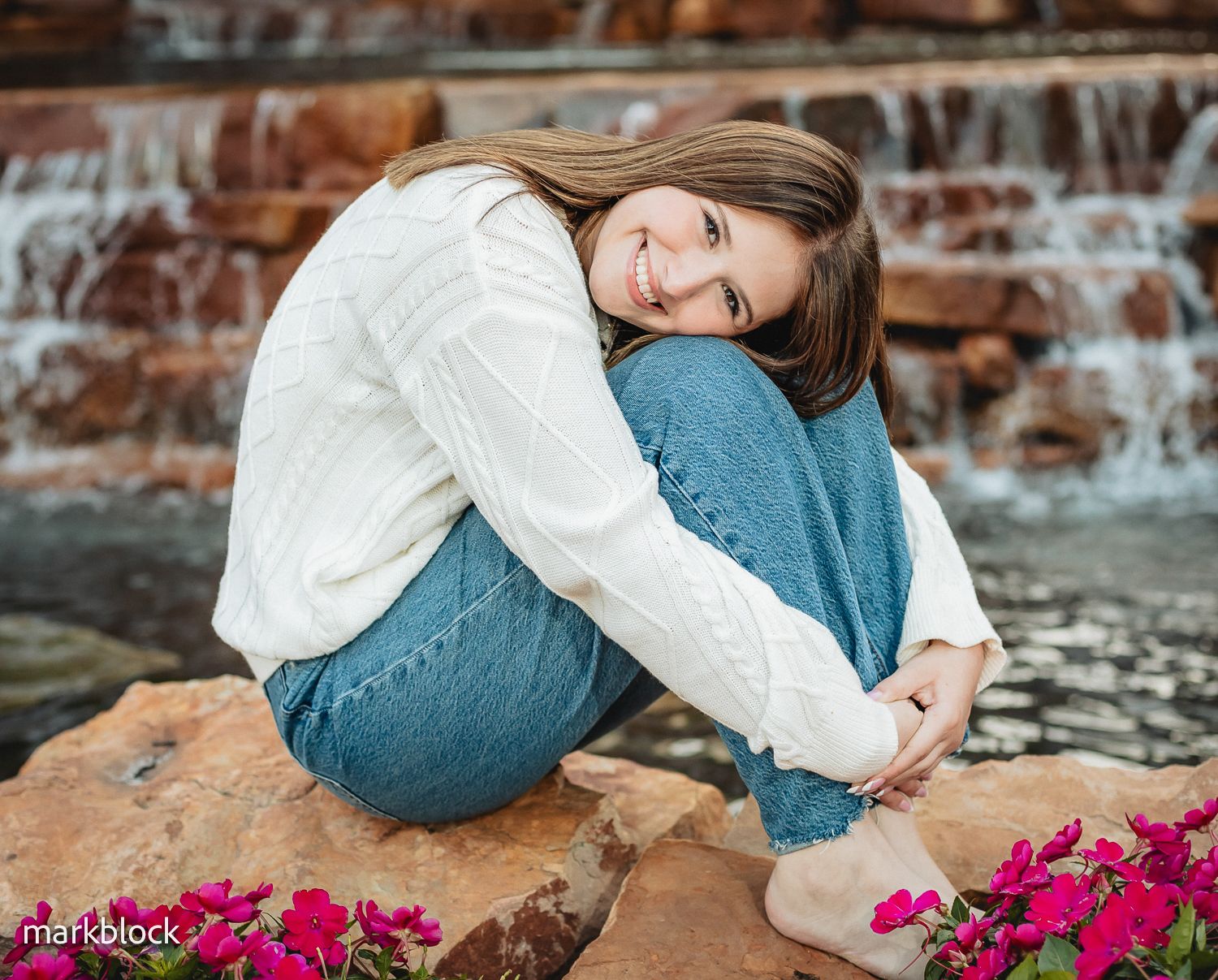 Someone in a white sweater and blue jeans sits on a rock near a waterfall with pink flowers in the foreground.