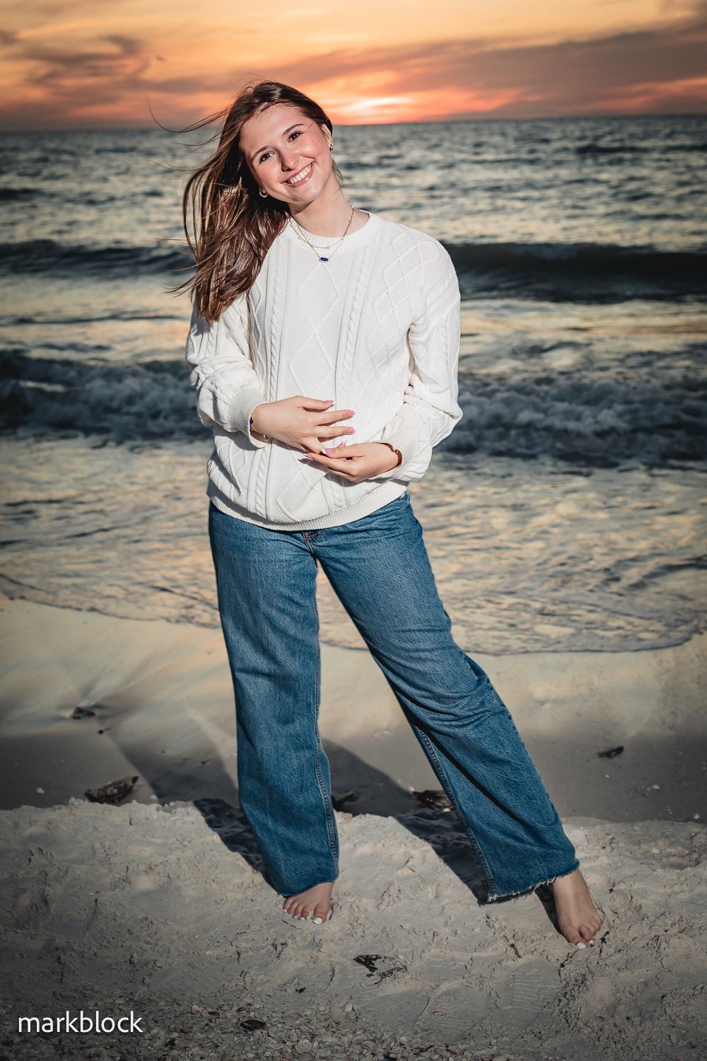 A beachgoer in a white sweater and jeans stands barefoot on sandy shores at sunset with waves crashing behind.