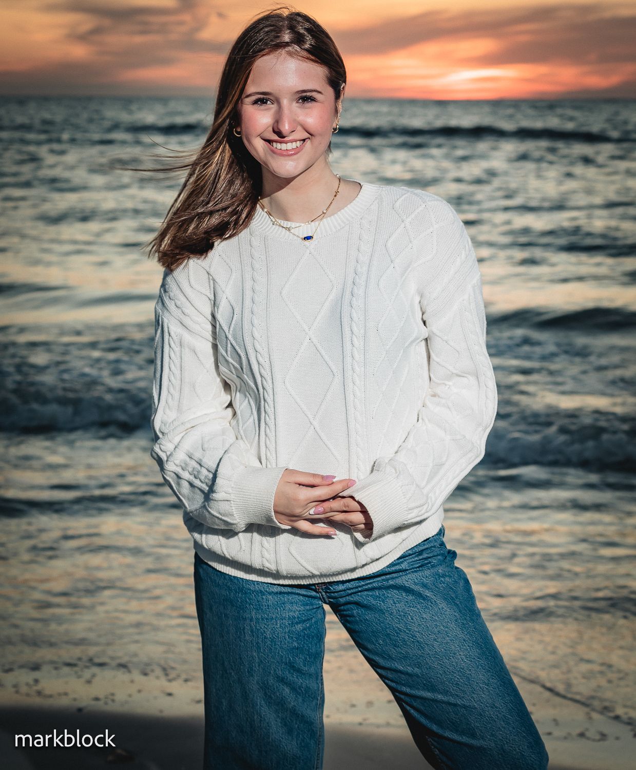 A person in a white cable knit sweater stands on a beach at sunset with waves and orange sky in the background.