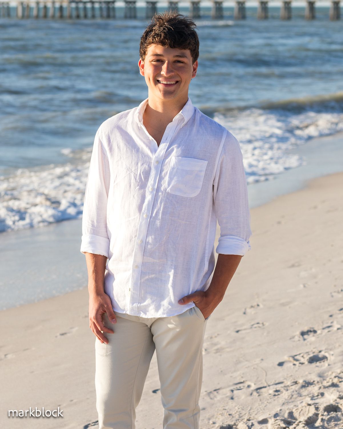 A person wearing a white button-down shirt and khaki pants stands smiling on a sunny beach with a pier in the background.