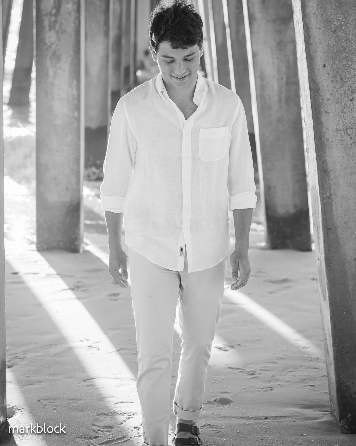Black and white portrait of someone wearing an all-white outfit walking under a pier with sunlight streaming through pillars.