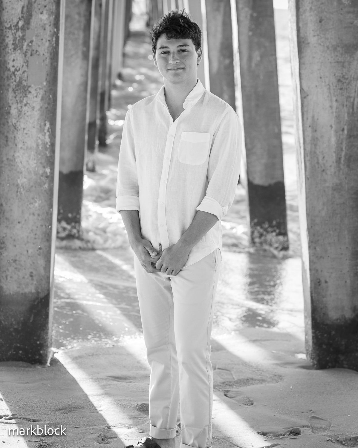 Black and white portrait taken under a pier showing a person in all white clothing standing between concrete pillars.
