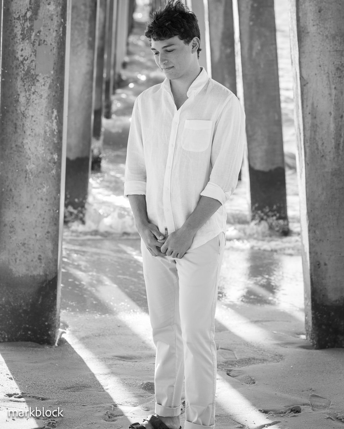 Artistic black and white portrait showing person in all-white outfit standing between concrete pillars with dramatic lighting.