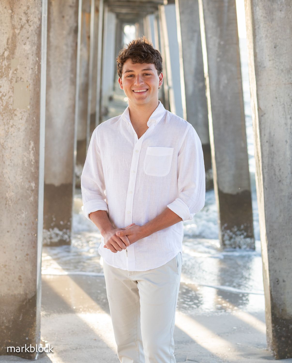 Someone in a crisp white button-down shirt and light pants poses under a pier with sunlight streaming between pillars.