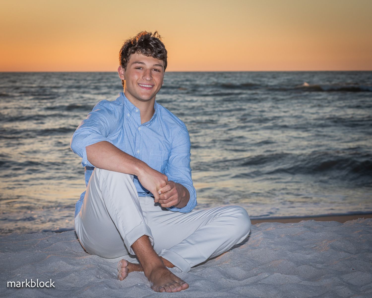 Sunset beach portrait of someone in a blue shirt and white pants sitting on sandy shoreline with ocean waves in background.