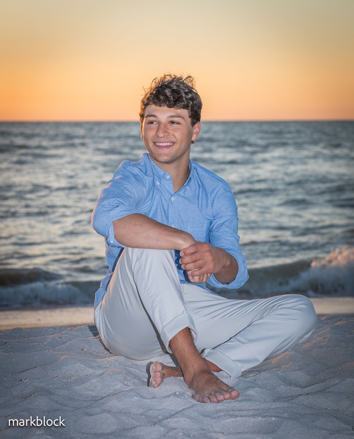 A person in a blue shirt and light pants sits on a sandy beach at sunset with the ocean waves in the background.