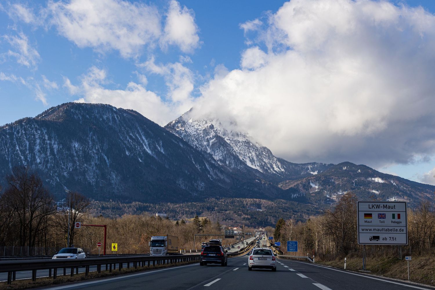A scenic mountain highway with snow-capped peaks and puffy clouds in the background.