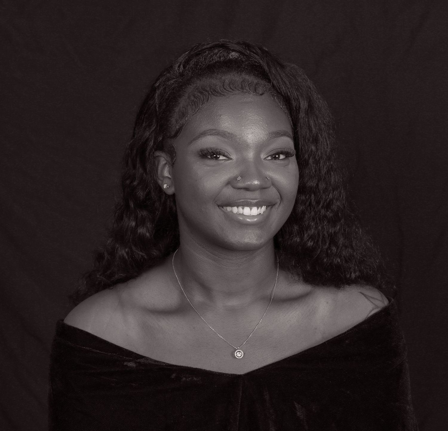 Beautiful portrait of a person with long curly hair wearing an off-shoulder black top and small necklace against a dark background.