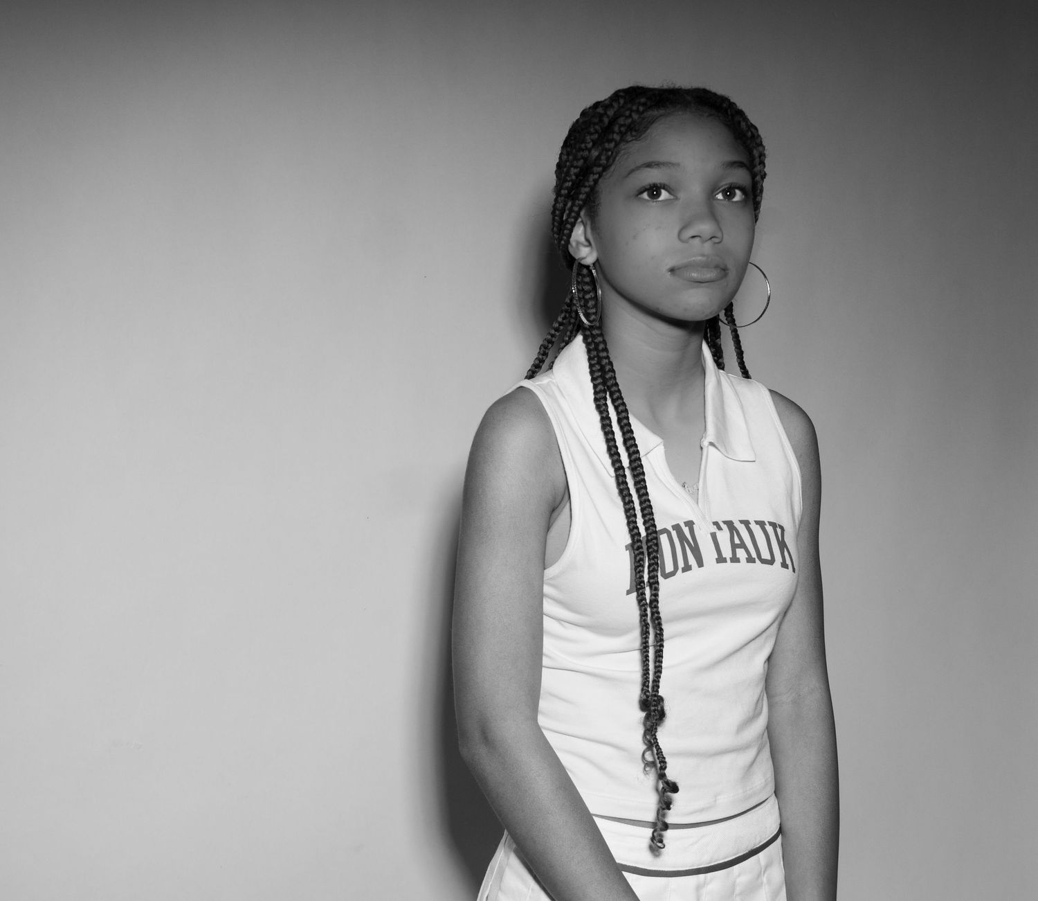 Person wearing white tank top with braided hair poses against wall in black and white portrait photography.