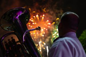 Military musician in uniform watching fireworks explode in the night sky, euphonium reflecting the vibrant colors.