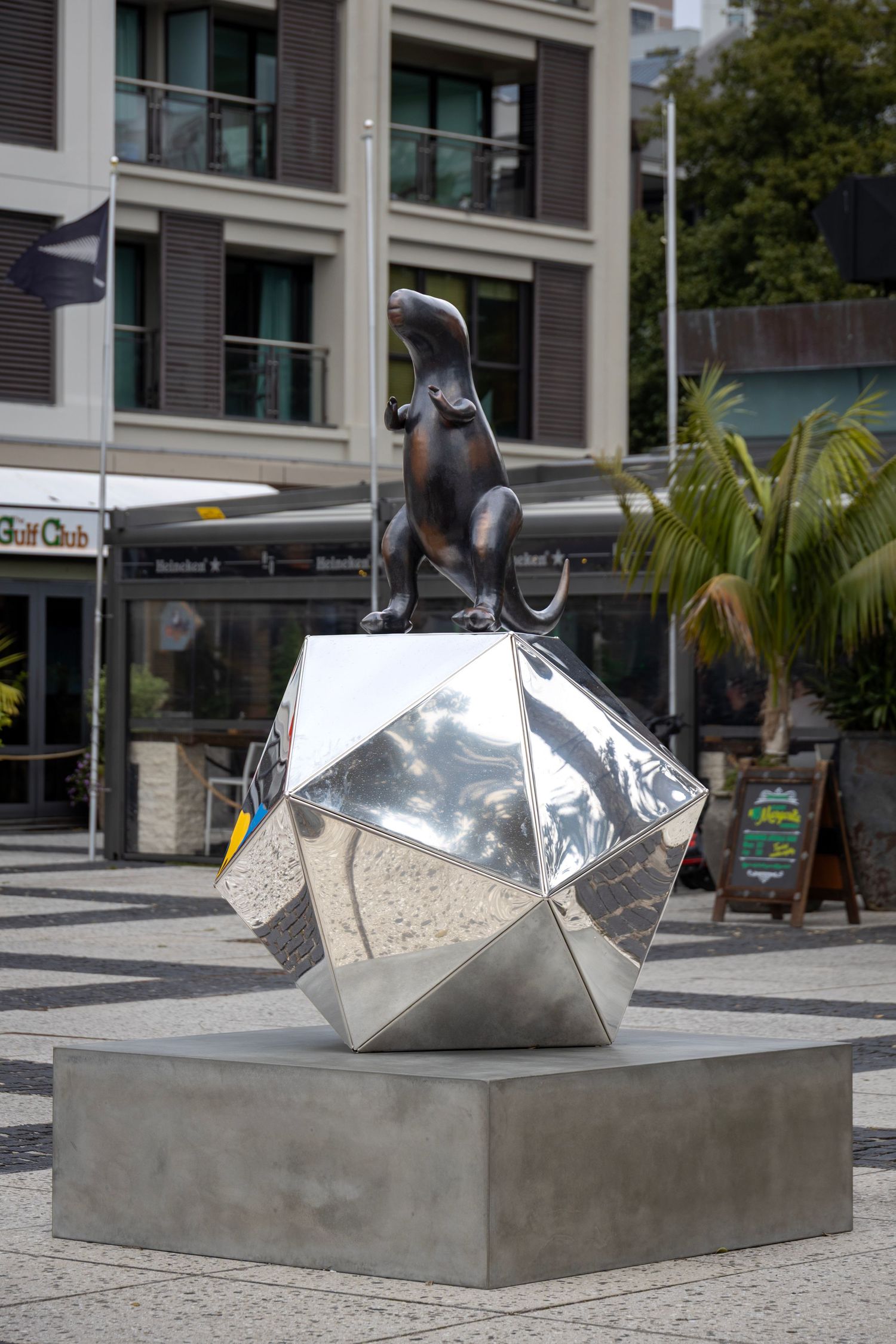Modern sculpture of a seated t-rex figure atop a faceted silver geometric base in an urban plaza with palm trees and buildings nearby.
