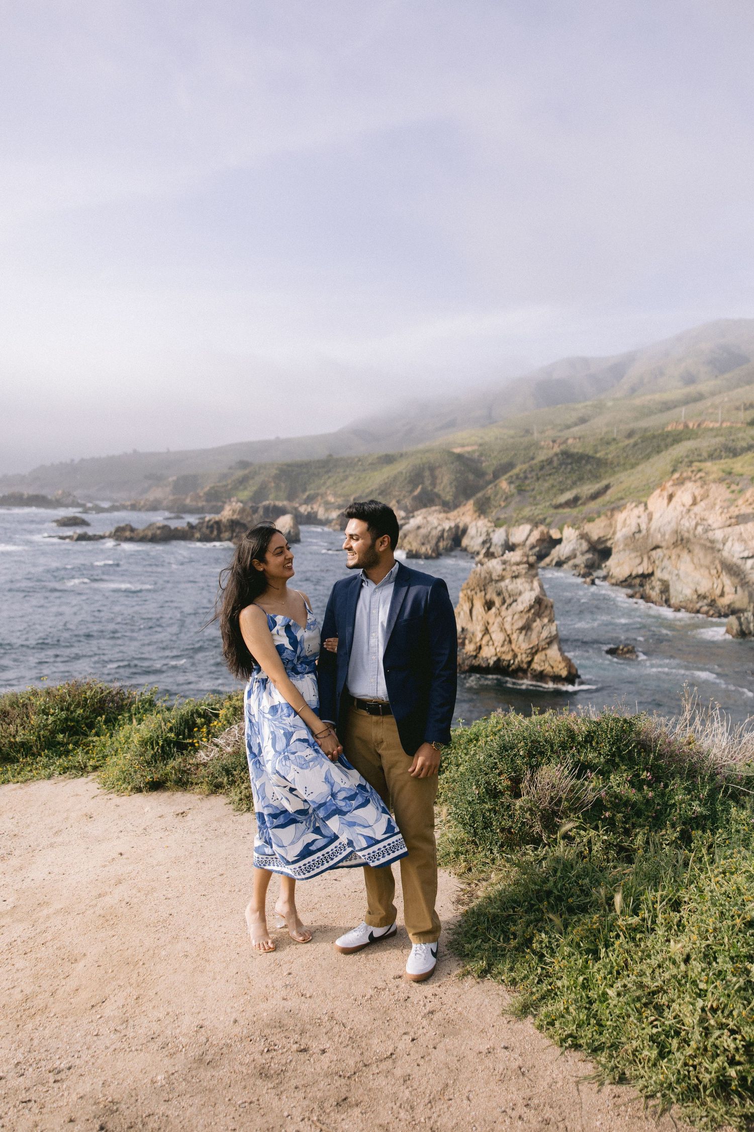 A couple enjoys a romantic moment along the rugged California coastline with cliffs and ocean views on a misty day.