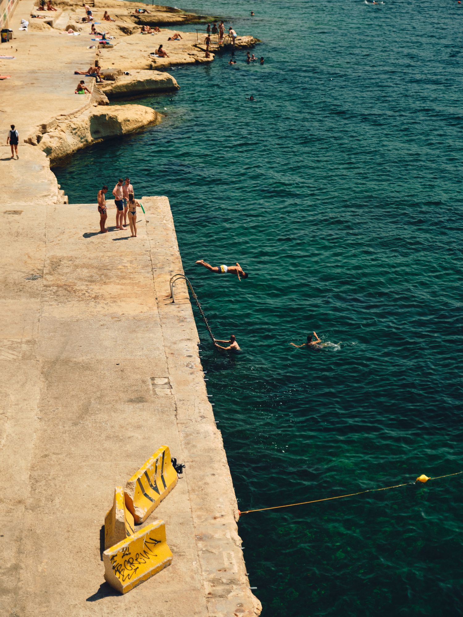 People swimming and sunbathing along a rocky Mediterranean coastline with turquoise waters and a yellow caution sign.