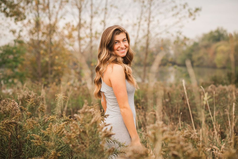 A person in orange top poses outdoors in natural lighting among tall grass.