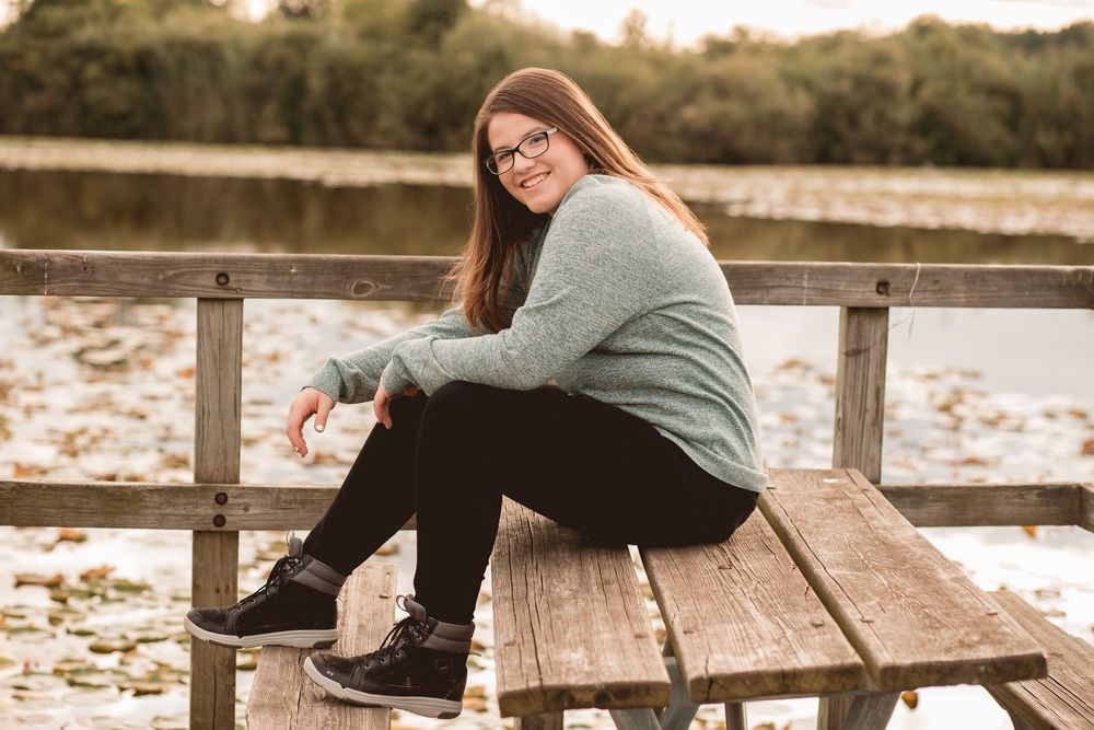 A person in casual wear sits thoughtfully on a wooden dock overlooking a lake.