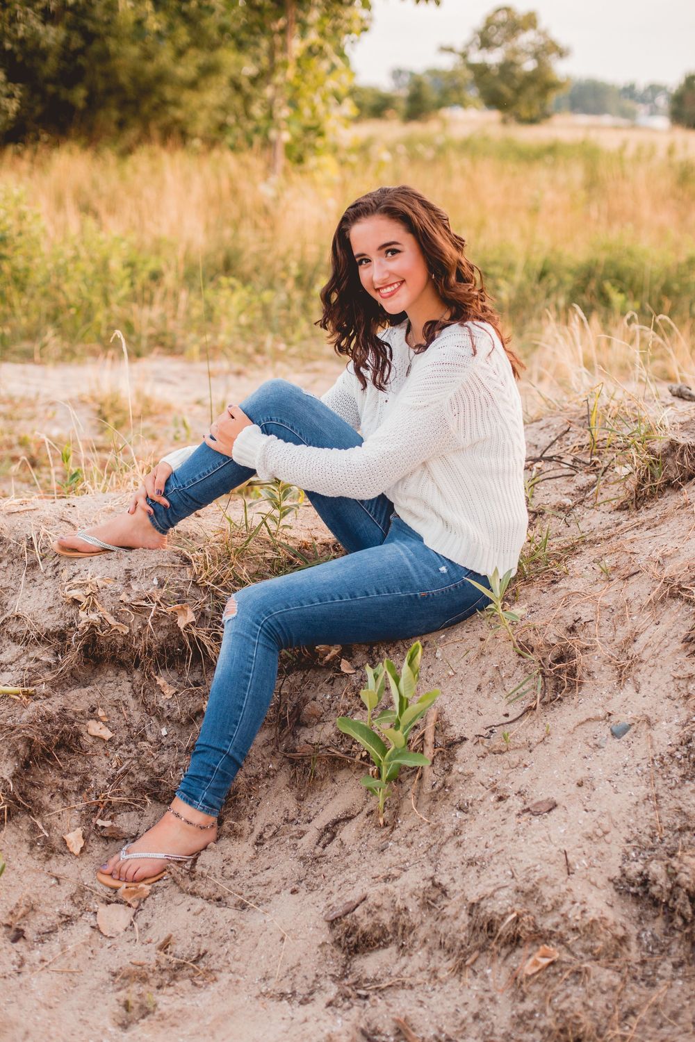 A person in jeans and white sweater poses on sandy ground during sunset.