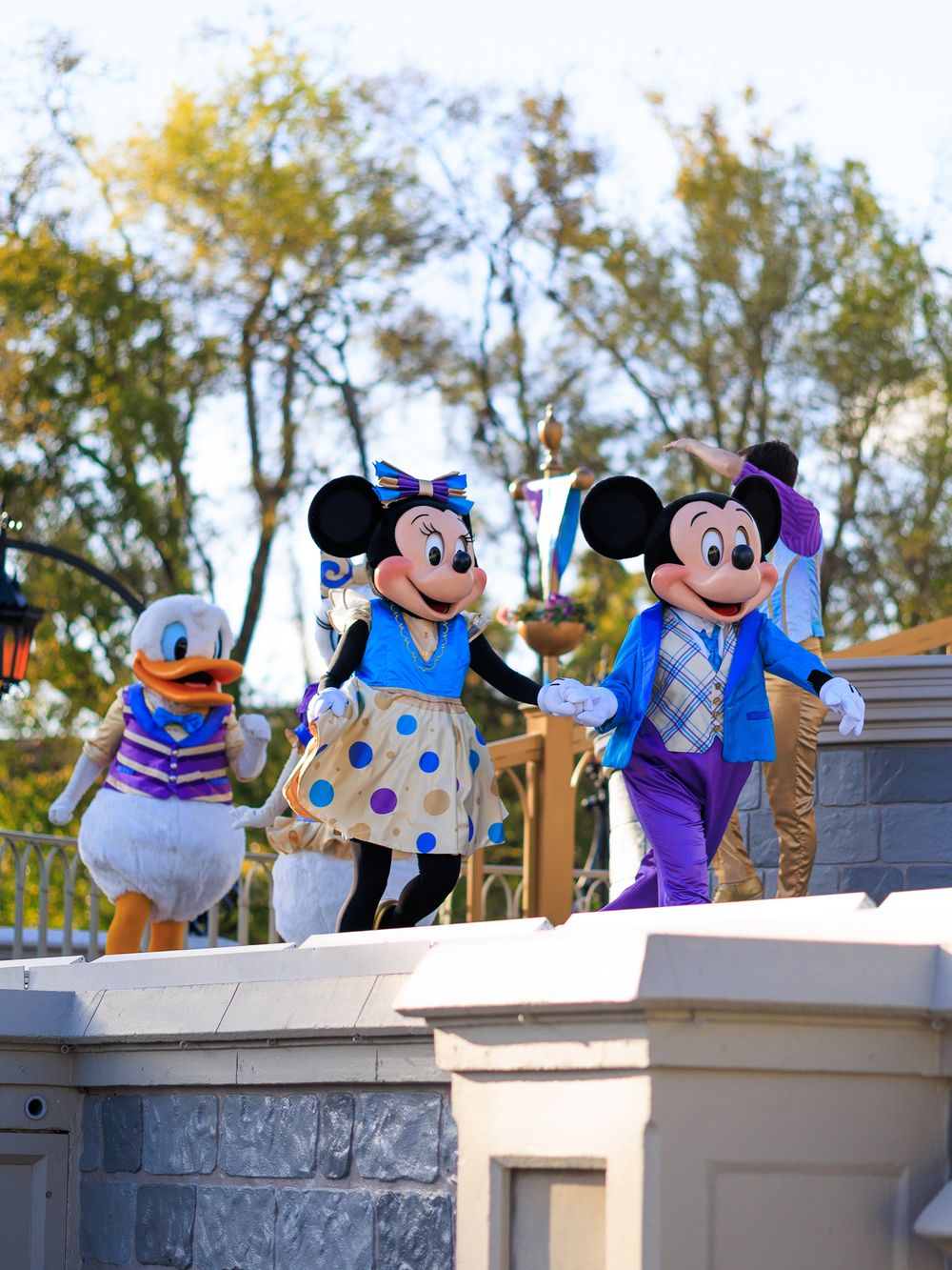 Disney characters Mickey Mouse, Minnie Mouse, and Donald Duck wave from atop a stone wall at Magic Kingdom.
