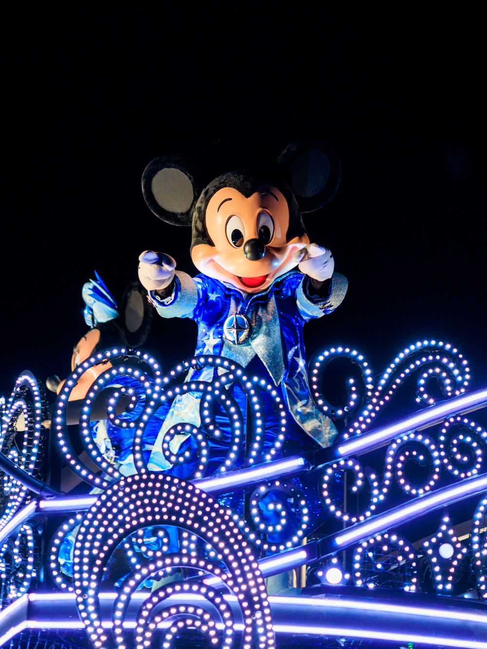 Mickey Mouse waves from a glowing blue illuminated float during Disney's nighttime parade spectacular.