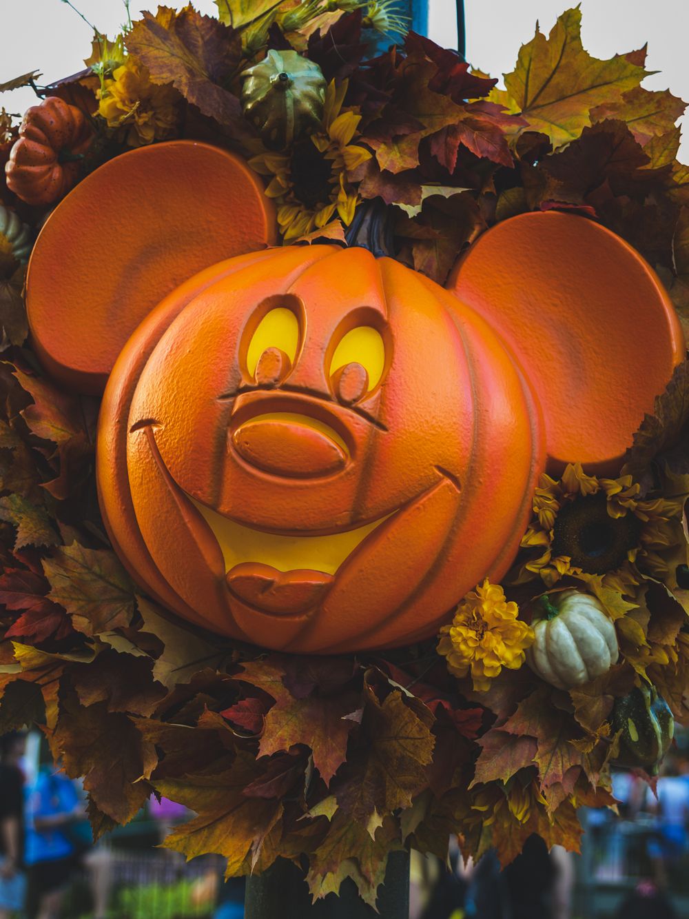 Smiling Mickey Mouse-shaped pumpkin decoration surrounded by fall foliage.