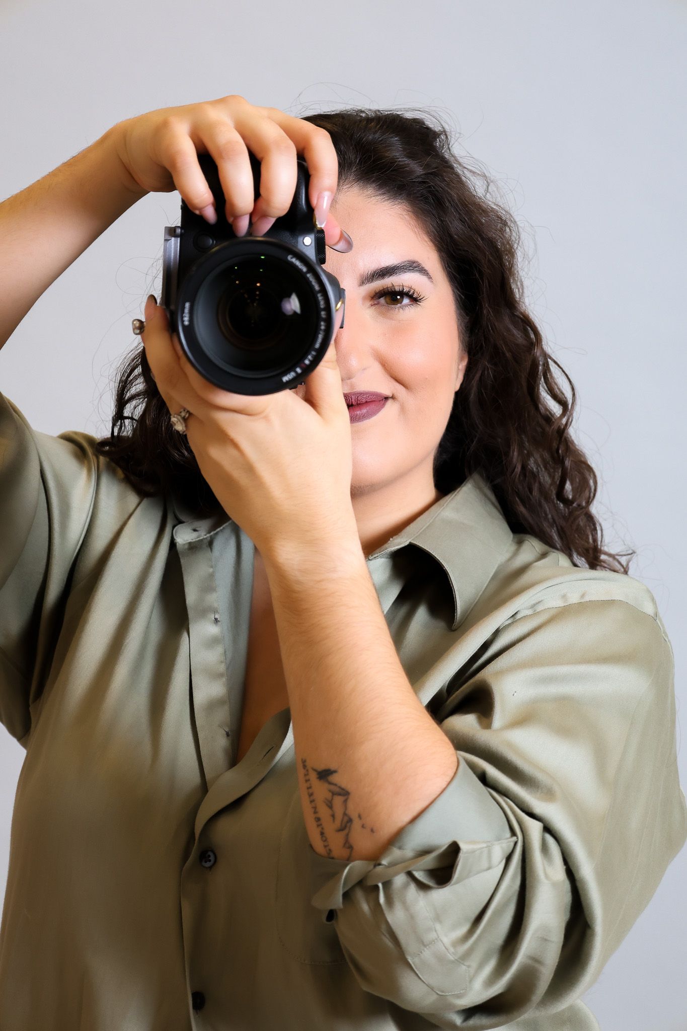 Photographer wearing sage green button-up shirt holds professional DSLR camera against white background.