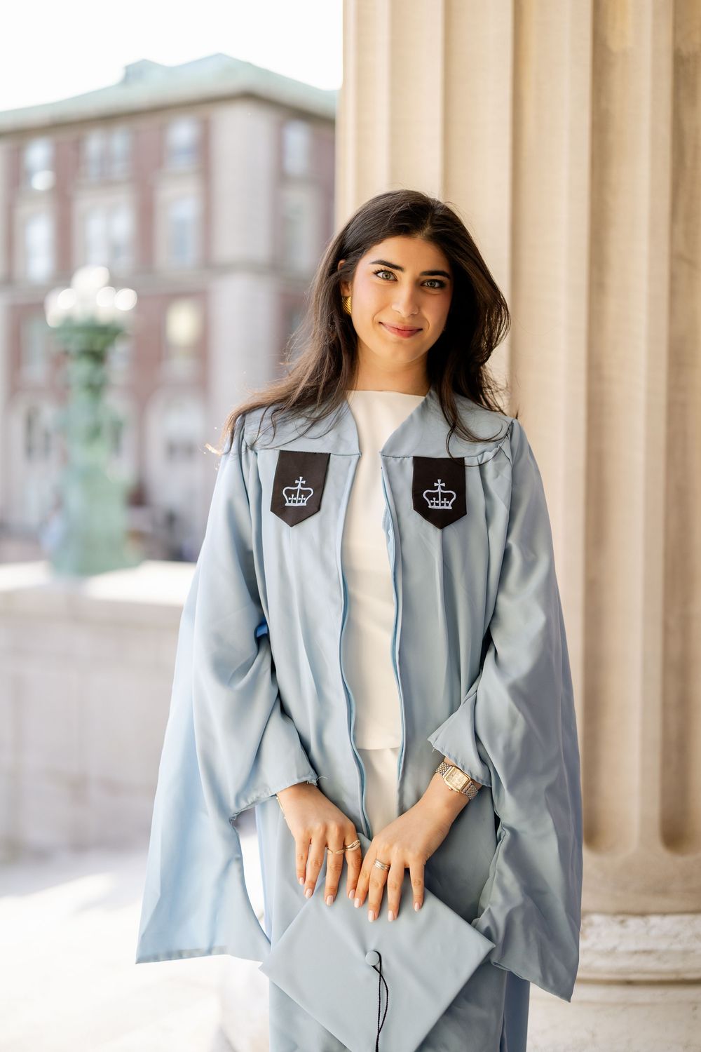 Graduate in light blue academic regalia with crown emblems poses by pillars at university campus.